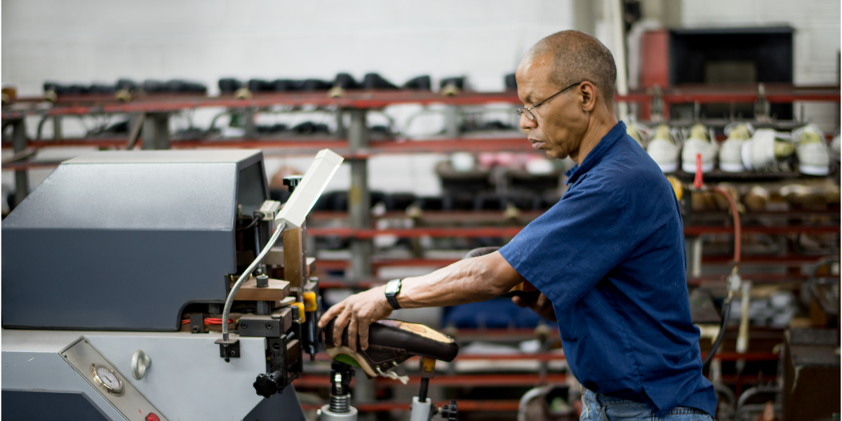Worker in glasses and a blue collared shirt using a stitching machine to finish a shoe, with racks of black and white footwear in the background.