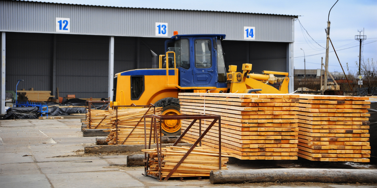 Front loader moving stacked timber planks in the yard outside a wood processing warehouse.