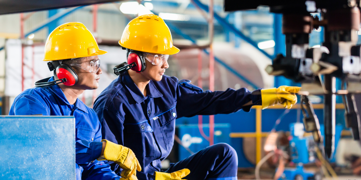 Two workers in hard hats, safety glasses and ear protection on a factory floor, one pointing towards overhead machinery near a crane hook.
