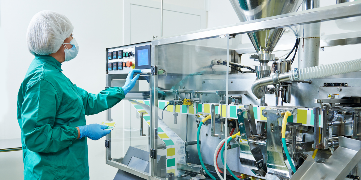 Worker in a hairnet, mask and gloves operating a stainless steel packaging machine in a clean manufacturing facility.